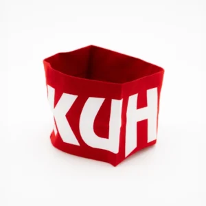 Auf weißem Untergrund steht ein roter, flexibler Behälter mit den Teilbuchstaben „KUH“ in Weiß, möglicherweise eine Anspielung auf das Stirnband „Kuhstall“ rot aus dem Kitzloch Shop.