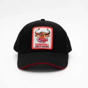 Die „Schildkappe Patch ‚Kuhstall‘ schwarz“, die im Kuhstall Kitzloch Shop erhältlich ist, ist eine schwarze Kappe mit rot umrandeter Krempe, mit einem Aufnäher in Form eines Bullen im Cartoon-Stil und dem Text „Kohstall Home of Warmen“ auf der Vorderseite vor einem schlichten weißen Hintergrund.