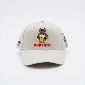 Die Schildkappe „30 Jahre Kuhstall“ ist eine beige Baseballkappe mit einem aufgestickten Cartoon-Stier, der einen Bierkrug hält, über dem Wort „KUHSTALL“ in Rot, das die spielerische Atmosphäre des Kuhstall Kitzloch Shops hervorruft. Die Kappe ist auf einem schlichten weißen Hintergrund abgesetzt.