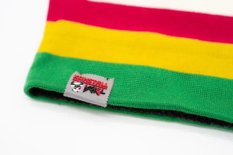 Die Mütze „Stripes“ grün rot gelb ist eine Strickmütze mit horizontalen Streifen in Rot, Gelb, Grün und Weiß. Sie wurde exklusiv für den Kuhstall Kitzloch Shop entworfen, hat ein kleines Etikett mit Text und einer Grafik auf der Krempe und wird flach auf weißem Hintergrund präsentiert.