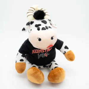 Die Plüschkuh, eine Kuschelkuh mit schwarzen und weißen Flecken, kleinen Hörnern und orange-braunen Hufen, ist mit einem T-Shirt vom Kuhstall Kitzloch geschmückt. Sie lehnt auf einem schlichten weißen Hintergrund, und ihr schwarzes Fellbüschel verleiht ihr einen charmanten, gemütlichen Look.