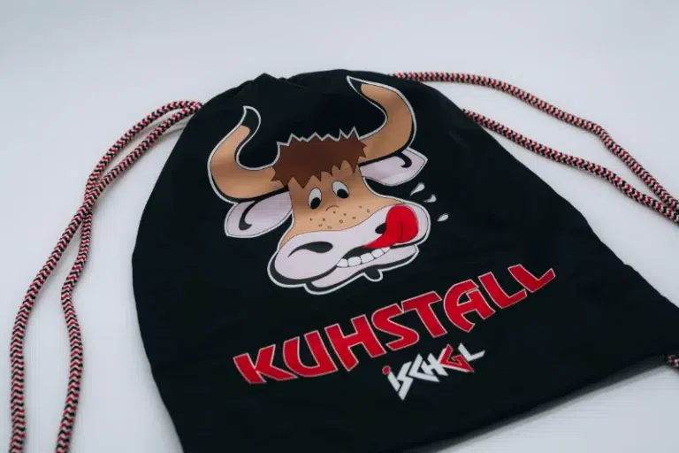 Der Kuhstall Rucksack, erhältlich im Kuhstall Kitzloch Shop, ist ein schwarzer Kordelzugbeutel, der mit einer verspielten Cartoon-Kuh verziert ist, die ihre Zunge herausstreckt. Unter der Kuh steht prominent in Rot „KUHSTALL“ mit einer stilisierten weißen Signatur darunter.