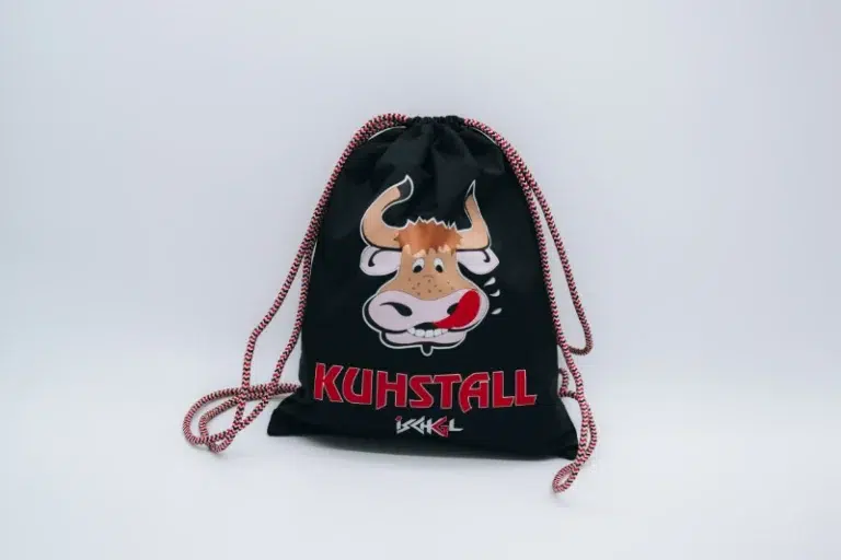 Der Kuhstall Rucksack, erhältlich im Kuhstall Kitzloch Shop, ist ein schwarzer Kordelzugbeutel, der mit einer verspielten Cartoon-Kuh verziert ist, die ihre Zunge herausstreckt. Unter der Kuh steht prominent in Rot „KUHSTALL“ mit einer stilisierten weißen Signatur darunter.