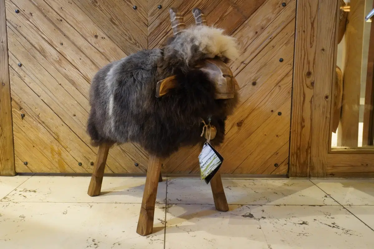 Das Fellhocker Schaf „Bua“ ist eine skurrile Schaffigur mit einem pelzigen grau-braunen Körper, der auf Holzbeinen steht. Es verfügt über einen bezaubernden Holzkopf mit kleinen Hörnern und einem Schild, das vor diagonal gemusterten Holzwänden auf einem gefliesten Boden steht.