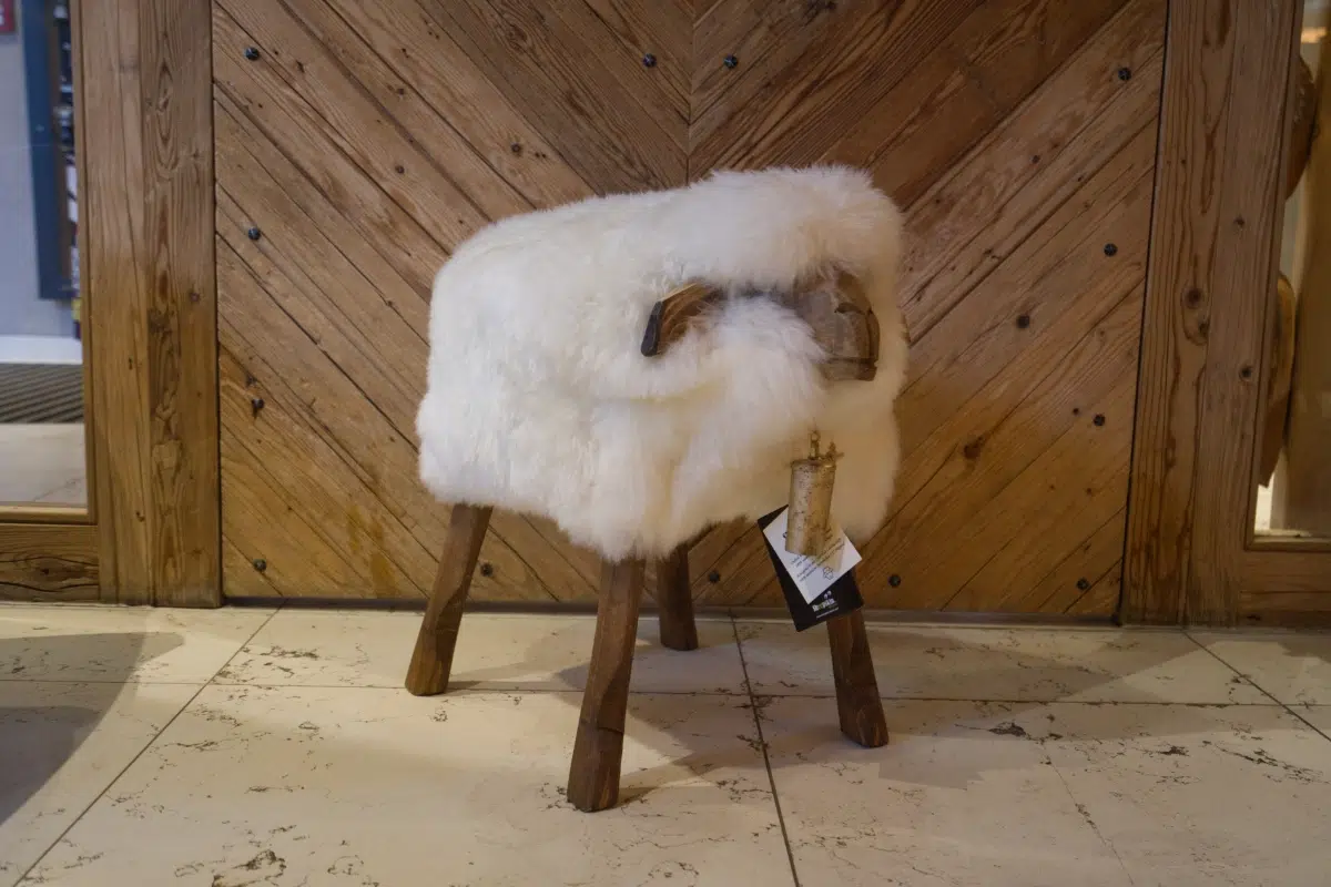 Der Fellhocker Schaf „Bua“ ist ein kleiner Hocker mit Holzbeinen, der mit flauschigem weißem Fell bedeckt ist und an ein Schaf erinnert. Er verfügt über gebogene Holzgriffe, die Schafshörner imitieren, und an dem Hocker hängt ein Preisschild. Er steht auf einem hellen Boden vor einer Holzwand.